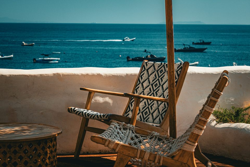 Fauteuils en bois tendus de toile tissée sur une terrasse à la chaux, la mer et des bateaux au fond